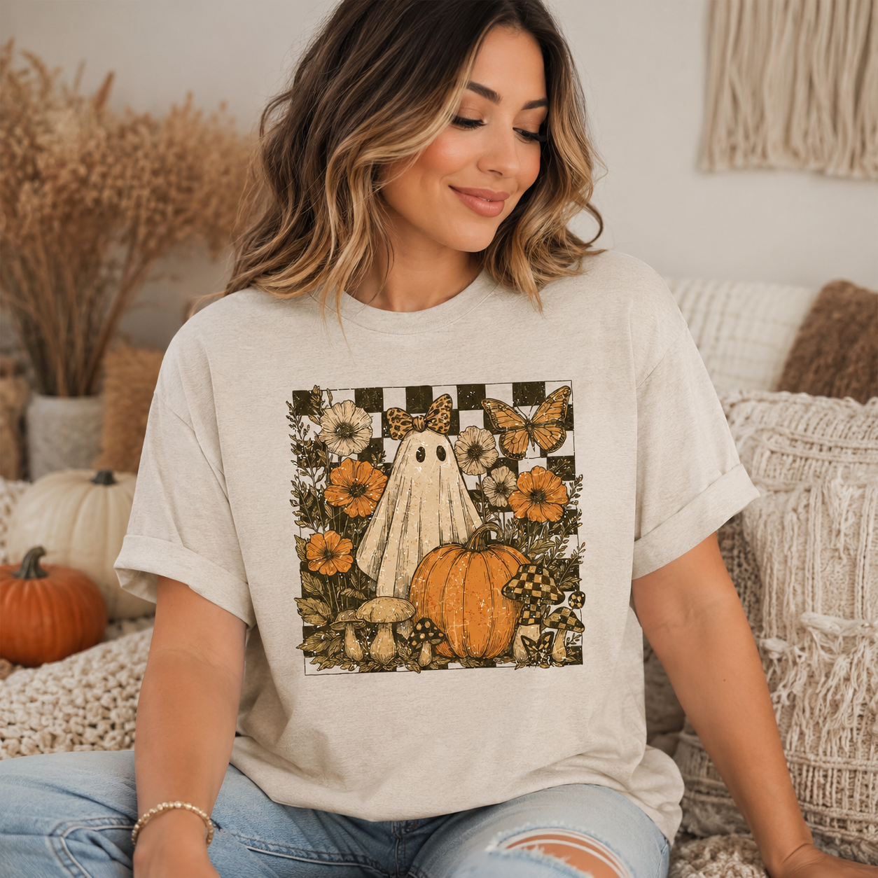 A woman wearing a light beige graphic t-shirt with a Halloween-themed print featuring a ghost, pumpkin, flowers, and butterflies, sitting on a cozy couch.