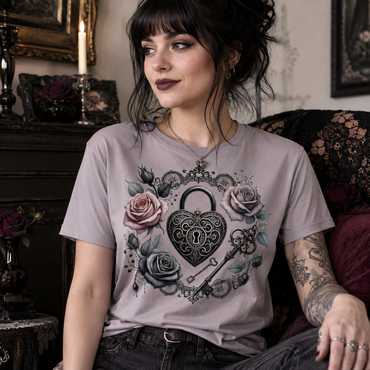 A woman wearing a light gray t-shirt with a gothic-style graphic of a heart-shaped padlock, roses, and keys, sitting in a vintage-style room.