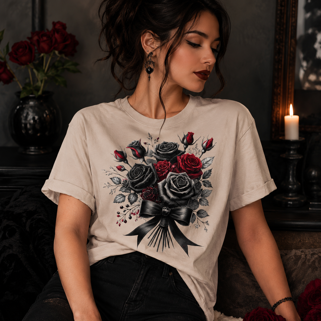 A woman wearing a beige graphic t-shirt featuring a black and red rose bouquet design, posed in a dark, moody room with candles and red roses in the background.