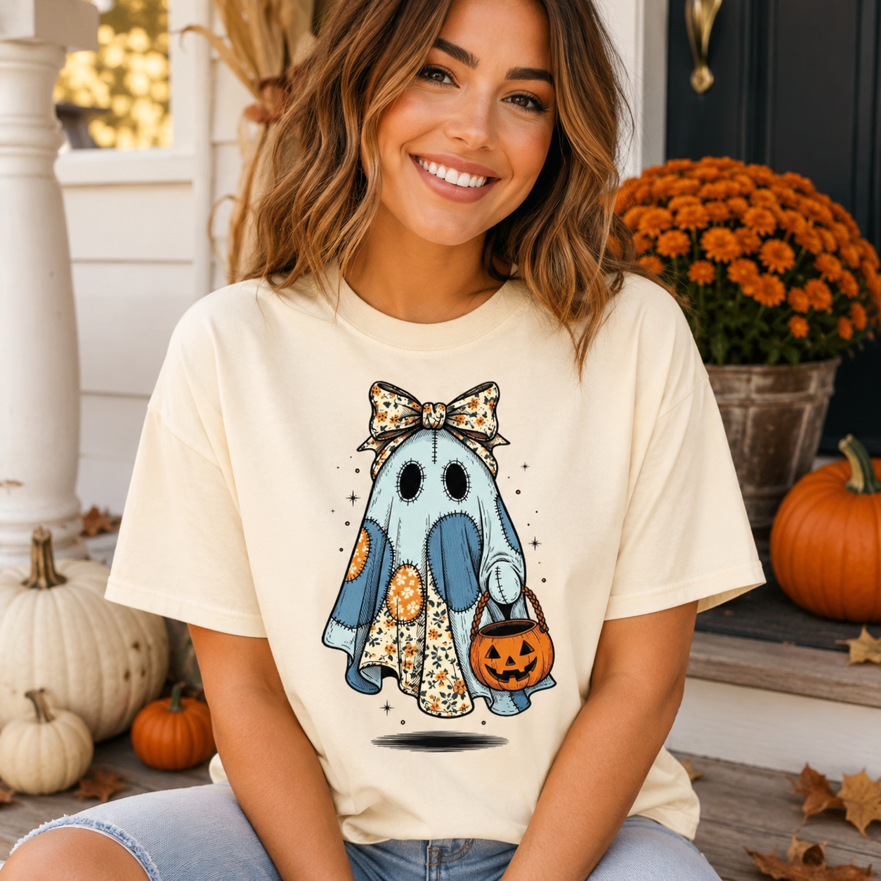 A woman wearing a cream-colored graphic t-shirt with a cute cartoon ghost Halloween design, posing on a porch decorated with pumpkins and orange mums.