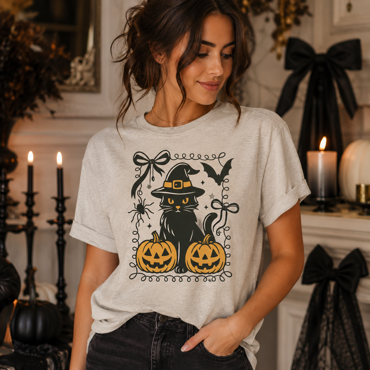 A woman wearing a light gray t-shirt with a black cat wearing a witch hat, sitting between two jack-o'-lantern pumpkins, and surrounded by Halloween-themed illustrations. The background shows a cozy, decorated indoor setting with candles and black bows.