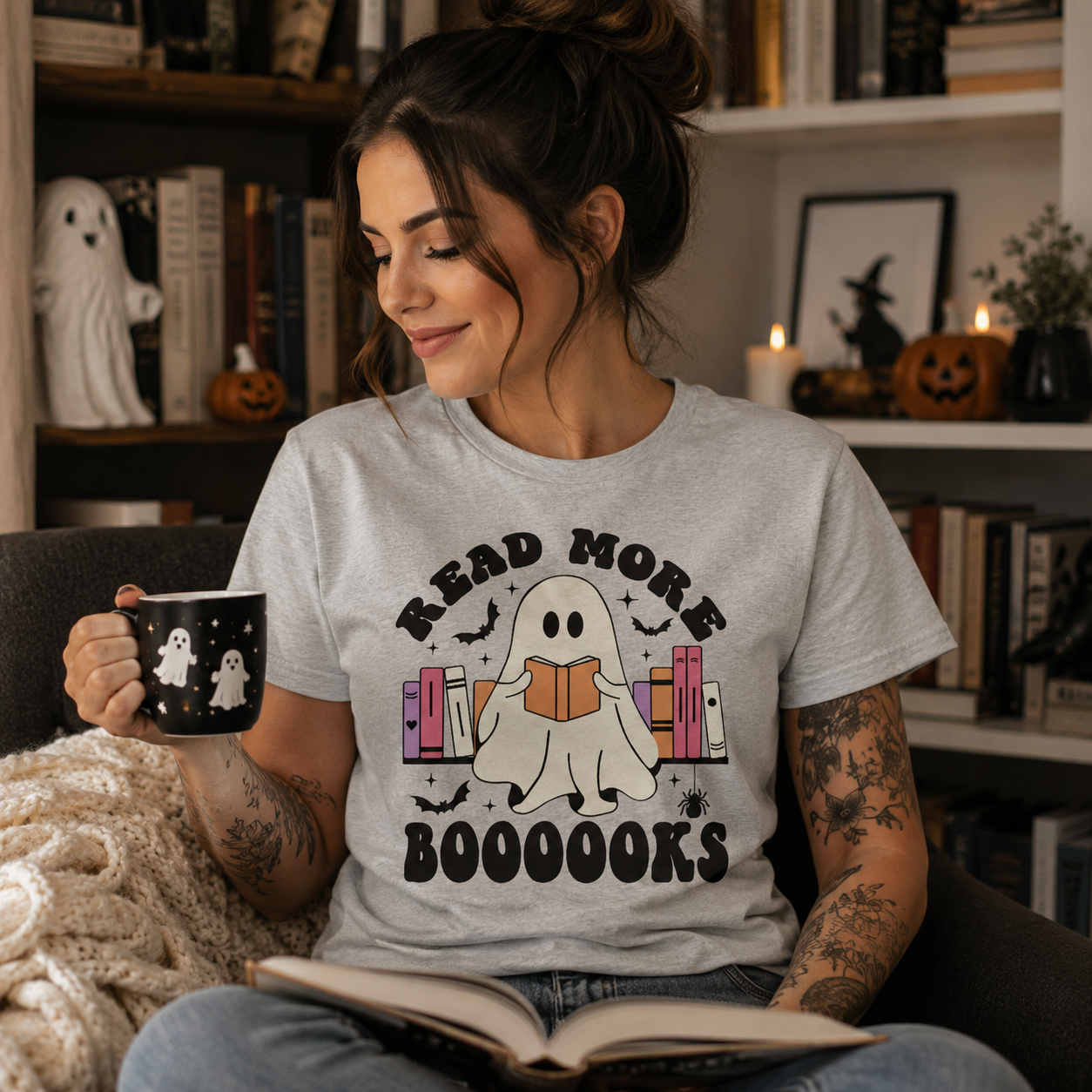 A woman sitting in a cozy room holding a mug and reading a book, wearing a light gray graphic t-shirt with a ghost and books design and the text 'READ MORE BOOOOOOKS'.