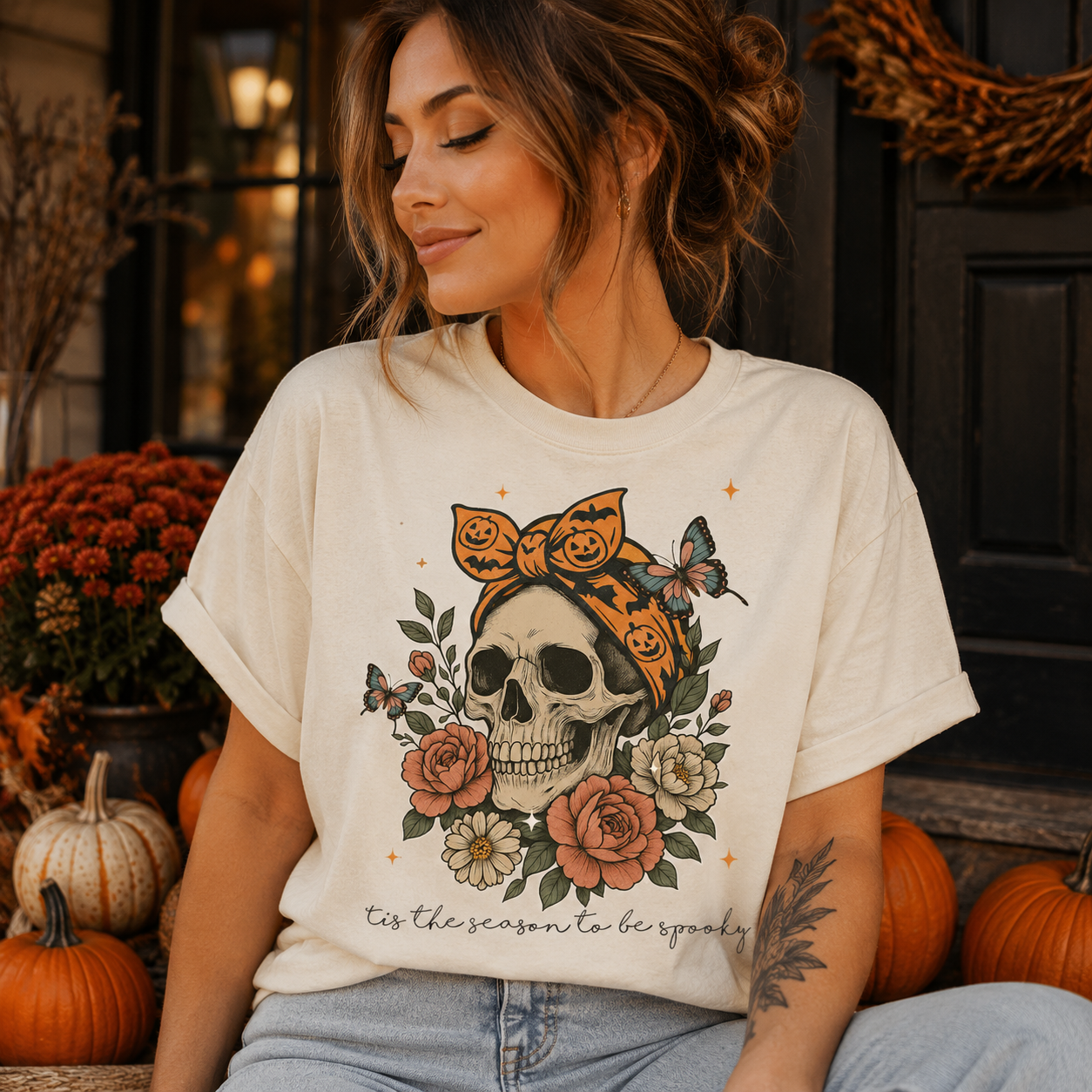 A woman wearing a cream-colored graphic t-shirt with a skull, flowers, and a bandana design, posed in an autumn-themed setting with pumpkins and orange mums.