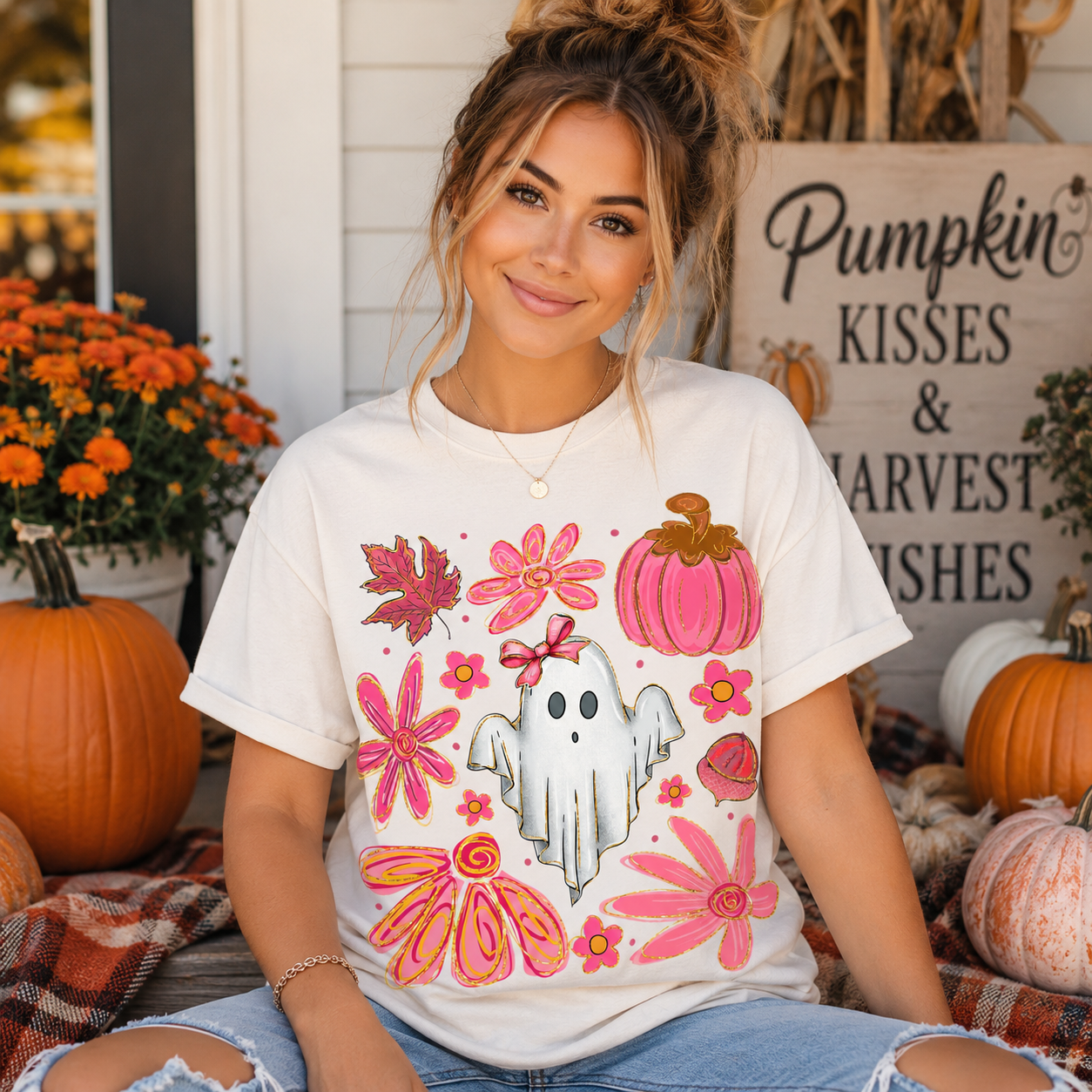 A woman wearing a white graphic t-shirt with a pink and orange autumn design, sitting in a fall-themed setting with pumpkins and a sign that reads 'Pumpkin Kisses & Harvest Wishes'.