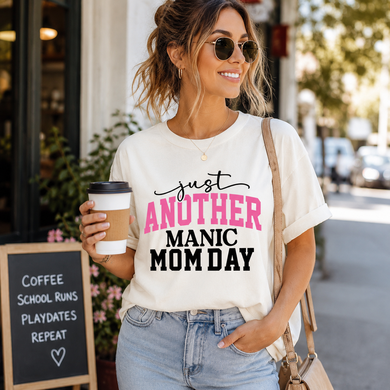 Woman wearing a t-shirt with 'Just Another Manic Mom Day' text, holding a coffee cup on a city street.