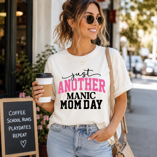 Woman wearing a t-shirt with 'Just Another Manic Mom Day' text, holding a coffee cup on a city street.